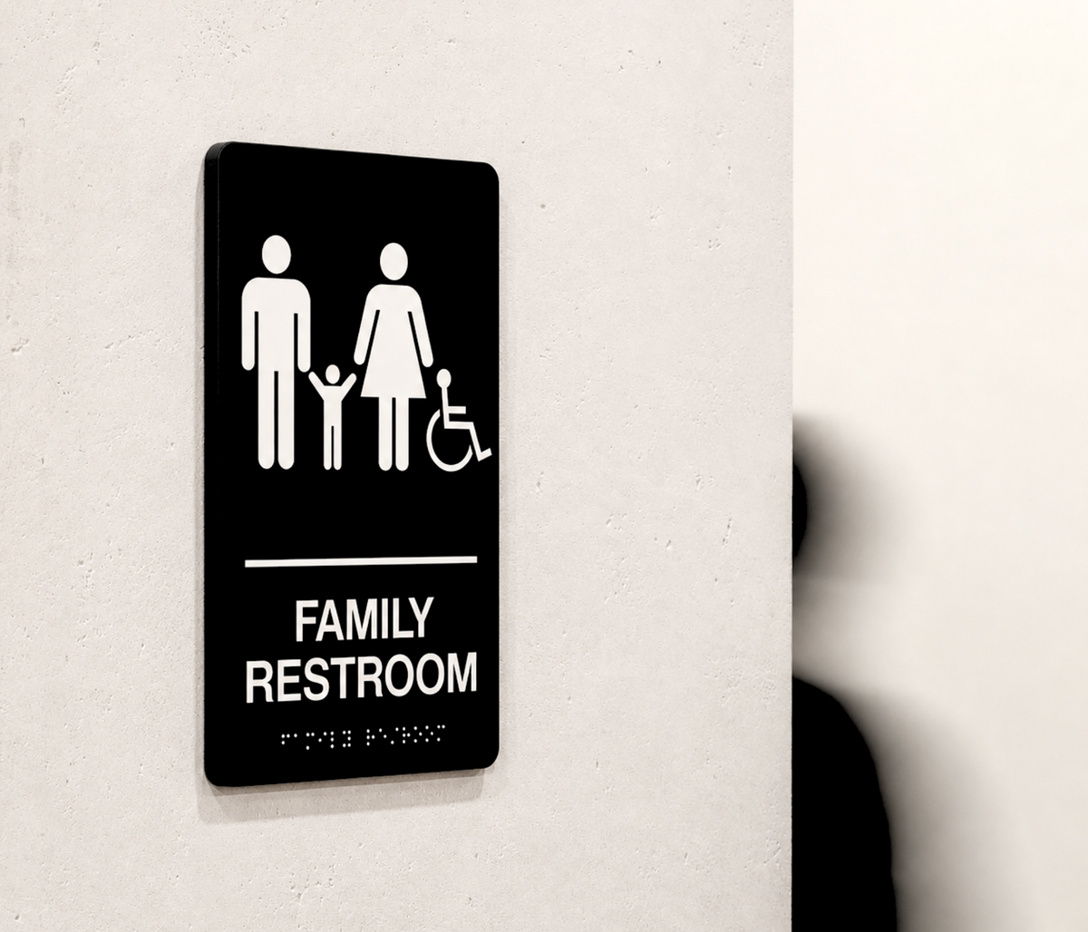 A black rectangular family restroom sign with white icons and text, mounted on a light beige wall. The sign shows a family and a wheelchair symbol, with the words 'FAMILY RESTROOM' and 'FOR THE PARENTS' in Braille below.
